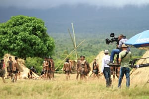Behind the scenes view of a film crew capturing a scene on an outdoor set with actors and natural scenery.
